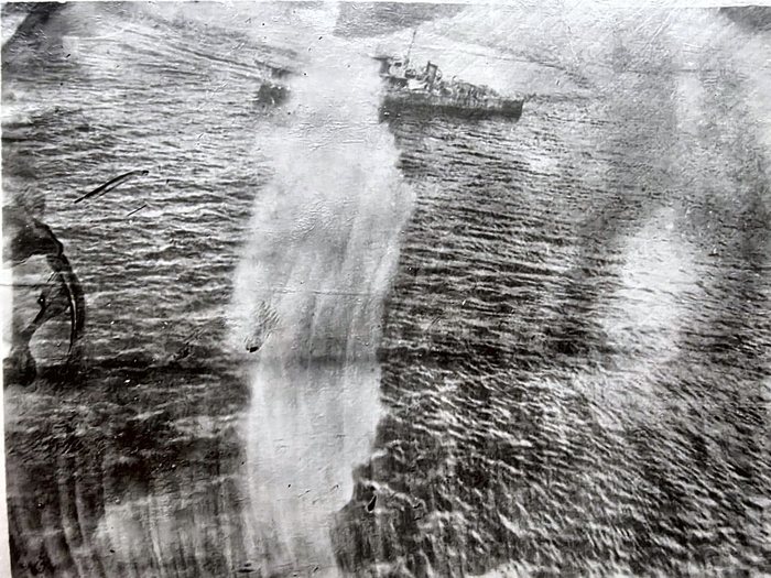A low oblique aerial photograph of a small warship under way, with broad pale plumes standing in the water around it and a wake trailing astern. The dark curved marks at the left edge are damage to the print.