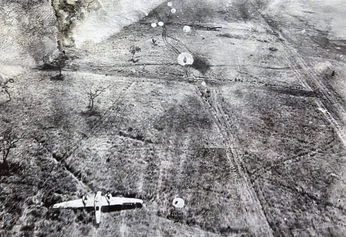 A near-vertical aerial view of open scrub laced with vehicle tracks: a twin-engine aircraft flies low across the lower part of the frame while small white parachute canopies descend over the ground and a column of smoke rolls up at the upper left.