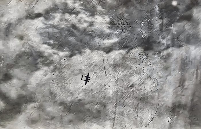 A twin-engine, twin-tailed bomber seen from above, dark against ground almost wholly veiled by smoke and blown dust, with scattered dark debris and a curving line of specks trailing off to its right.