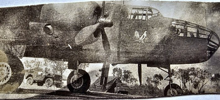 A B-25 Mitchell with its bombardier nose unmodified, parked at a dispersal, a white strutting cockerel painted on the starboard nose below the cockpit. A jeep with two men aboard, a tarpaulin shelter and a stake-bed truck stand in the sunlit background. Two other prints here show the same cockerel with EARLY BIRD lettered beneath it, so this is probably that aircraft; no name is painted below the bird in this frame, and no serial or unit marking is in view.