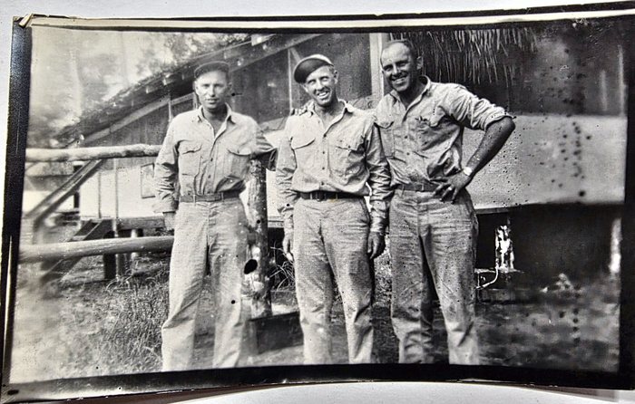 Three men in khaki shirts and trousers, two of them in caps, stand arm in arm in front of a raised hut with a thatched eave and a log railing; the back of the print carries three pencil signatures, too faint to read with any confidence, so none of the men is named here.