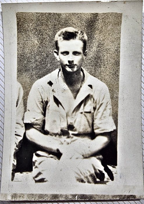 A young man in a light short-sleeved open-collar shirt sits cross-legged on the ground, hands resting in his lap, with the shoulder and knee of a second man just inside the left edge of the print. The back names him as Wilton M. Bunn of Troy, North Carolina -- the given name clear, the surname doubtful -- and carries a US Army censor stamp.