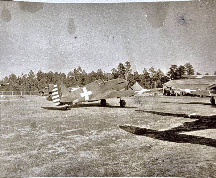 A Curtiss P-40 -- a single-engine low-wing fighter with an inline liquid-cooled engine and a tailwheel -- standing on a grass field, its fuselage and upper wing painted with large white crosses and its fin and rudder in broad stripes. A cargo truck is parked at the left behind the tail; at the right, in front of a long open-sided shed, stand a truck fitted with a boom and further aircraft carrying the same white markings, one of them cut by the edge of the print, with pine woods along the far edge. Its back is rubber-stamped 