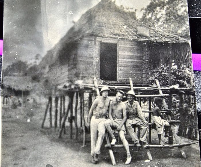 Three servicemen sit in a row along a heavy log rail in front of a New Guinean house raised on a forest of thin posts, with a sago-thatch roof and plank and woven walls; two of them are bare-chested and two wear wide sun helmets. At the right end of the rail a New Guinean child stands beside them, bare-chested and barefoot in a pale cloth wrap, turned away from the camera. A second house of the same build stands in the left background, fogged by a light leak. The back names the men in pencil -- 