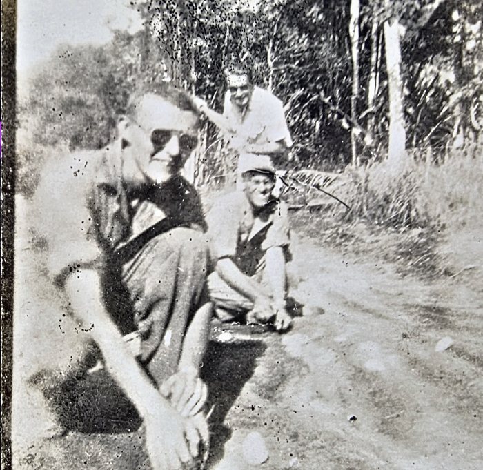 Three men in shirtsleeves on a dirt track at the edge of scrub bush: one kneels in the foreground in a dark shirt and cap, another crouches barehanded over something on the ground, and a third stands behind in a light shirt. The back gives three surnames with home states and the caption "Cracking Coconuts" -- "Sweigert Pa." is Allen naming himself, alongside "Davidson Ky." and a first surname read uncertainly as Funell, Furrell or Fennell.
