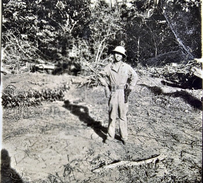 A serviceman in one-piece coveralls and a pith-style sun helmet stands hands-on-hips on cleared ground, a web belt at his waist with a dark holster at his left hip, felled timber and dense bush behind him. The back carries a single signature read as "Haskin[?]" -- Haskins or Hoskin are also possible -- with "N. G." for New Guinea, and the censor stamp "PASSED BY / BASE / 2415 / U.S. ARMY / EXAMINER."