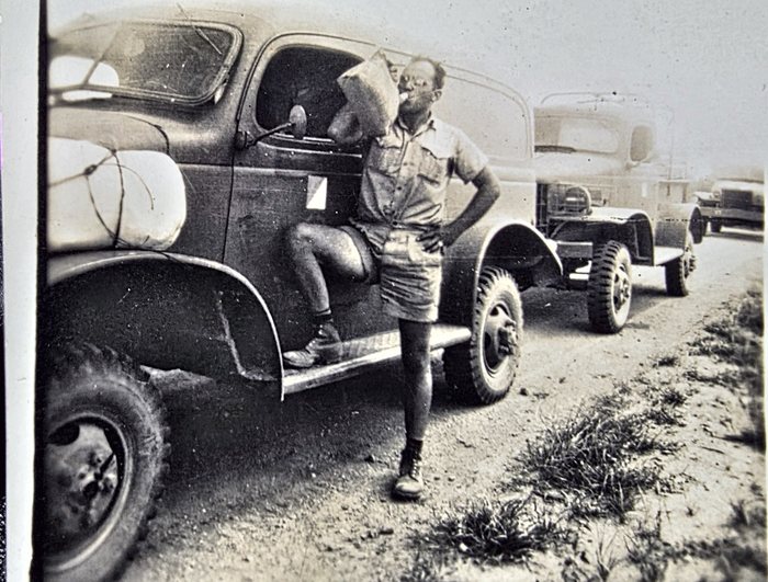 Allen in a work shirt and shorts, leaning against the closed steel cab of a military truck with one boot up on the running board and a cigarette in his mouth, a pale bundle lashed to the front fender and a line of further trucks stretching away down the dirt road behind. The back is a caption rather than a name: only the first word "Driving" and the closing line "in Australia." can be read, the middle left unresolved in pencil.