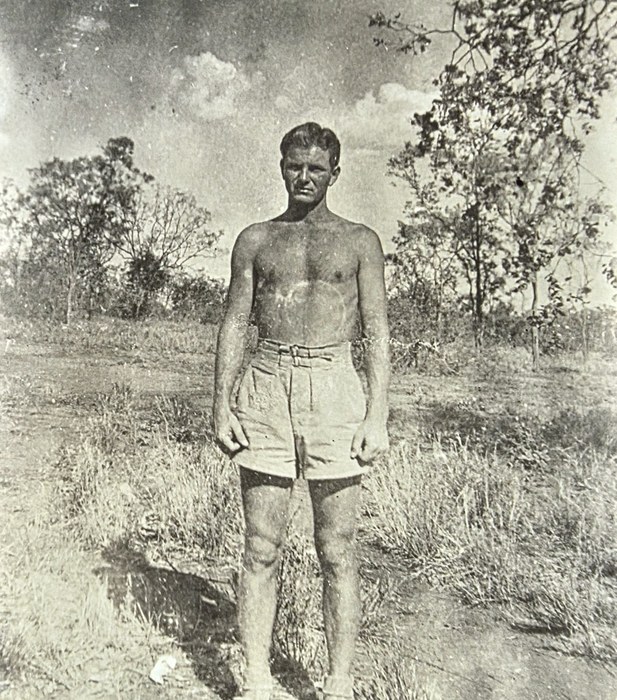 A bare-chested man in belted shorts stands squarely facing the camera on open, dry scrubland with thin trees behind him, the small print shown as it was photographed on a desk. The back reads "George E. Thom...", the surname running off the edge of the print after "Thom".
