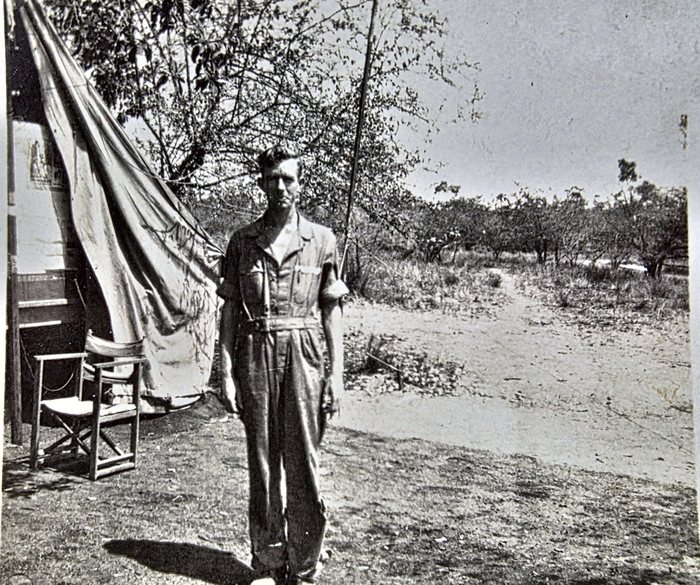 A serviceman in a one-piece work suit with the sleeves turned back stands bareheaded on bare ground beside a tent, its canvas wall drawn up and a folding wood-and-canvas camp chair set at the entrance, with open scrub and thin trees behind him. Named on the back as R. L. Thomas of Arlington, Massachusetts; the print also carries a US Army censor stamp.