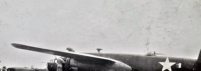 A narrow strip of a photograph showing a parked bomber from the side -- a wing running out to the left with its tip toward the camera, the framed cockpit glazing and a rounded engine cowling below it, and a large plain white star national insignia, with no bar, further aft on the fuselage. One man in light clothing stands at the wing root beside the engine, and a low treeline runs behind the aircraft. No name is written on the back; the reverse carries only the censor stamp "PASSED BY / BASE / 2415 / U.S. ARMY / EXAMINER".