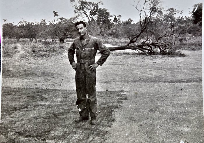 A serviceman in a dark one-piece work suit, belted at the waist, stands with his hands on his hips in open grassland, a fallen dead tree lying behind him and low scrub along the horizon. Named on the back as S. L. Taylor -- the middle initial could equally be an R -- with no home town given, and the reverse carries a US Army censor stamp struck twice.