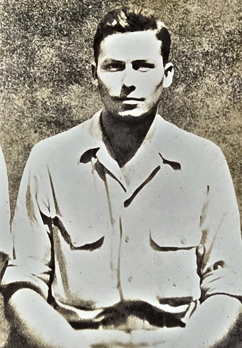 A young man sits with his hands loosely in his lap, wearing an open-collared light shirt with two flap breast pockets and the sleeves rolled above the elbow, photographed against a mottled backdrop. The name line on the back is clipped by the edge of the frame and cannot be read; beneath it the back gives "Westphalia, Kan", and a partial US Army censor stamp is present.