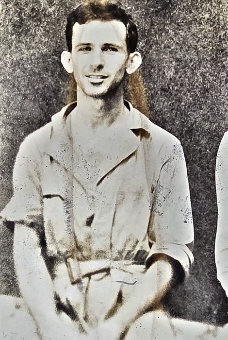 A young man sits smiling for the camera in a light open-necked shirt with breast pockets and turned-back sleeves, a belt at his waist and his hands folded in his lap, with the white sleeve of a second man at the right edge of the print. The back gives the surname as "Hoskins[?]" with the given name clipped by the frame, and beneath it "Galveston, Texas"; the censor stamp reads "PASSED BY / BASE / 2415 / U.S. ARMY / EXAMINER".