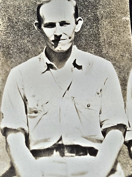 A thin-faced man sits in a light open-collar shirt with two buttoned breast pockets and a web belt at his waist, hands resting in his lap; another man's sleeve is just visible at the right edge. Named on the back as Henry Gosztyla of Hamtramck, Michigan.