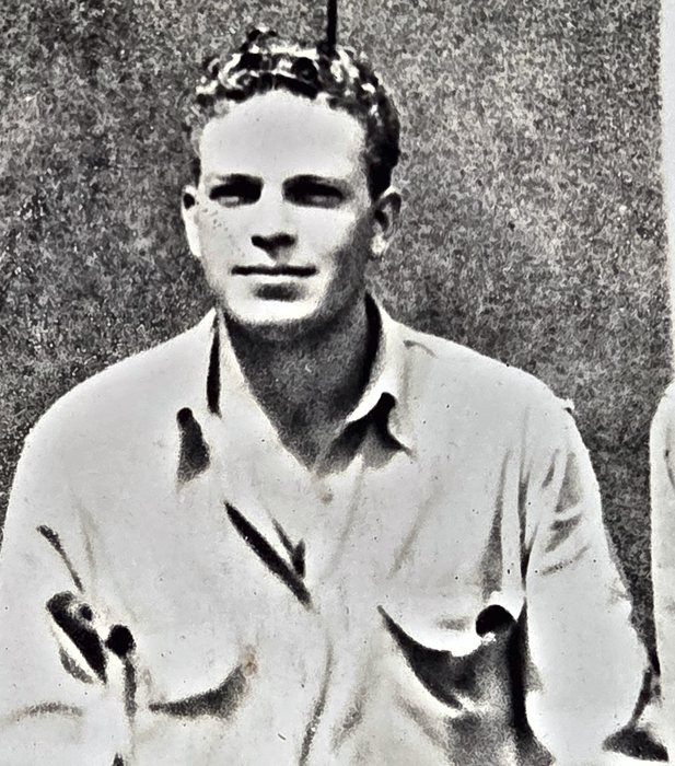 A wavy-haired man sits in a light open-collar shirt with two flapped breast pockets, half-smiling at the camera, the shoulder of the next man showing at the right edge. Named on the back as John B. Nunn of Springerville, Arizona.