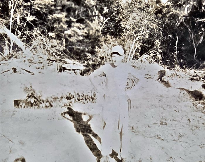 One man stands alone in a cleared patch of open ground, hands on his hips in light trousers and shirt and a soft cap, with scrub and trees close behind him. The back gives the surname Miller with "N. G." for New Guinea and the date "11/42"; the two readers of the handwriting differed on the given name, one reading "Hank" and the other "Herb".