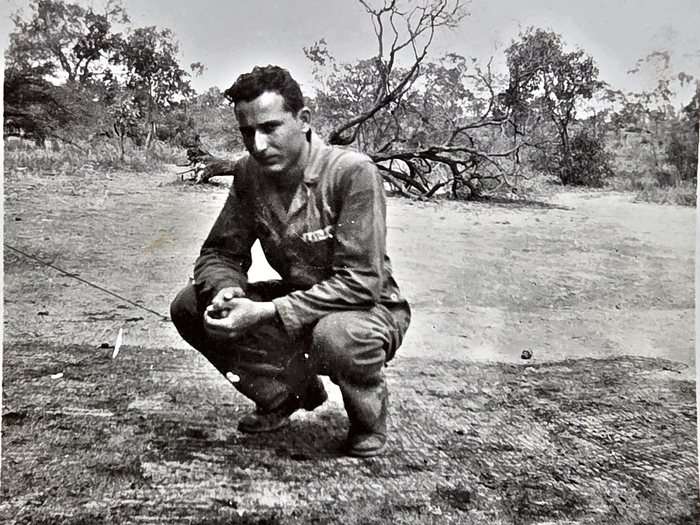 A man in a dark work shirt and trousers crouches on bare, dry ground among low scrub, a fallen bare-branched tree lying behind him, holding something small in his cupped right hand. The back of the print reads "L. L. Taylor" over "Los Angeles, Cal." -- the surname is certain, while the second initial was read as L by one reader and as J by the other.