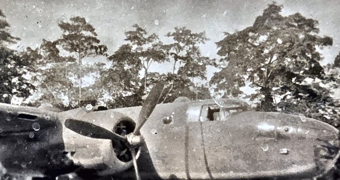 The nose, cockpit glazing and one three-bladed propeller of a bomber standing in a clearing at the edge of tall timber, with no one in the frame. The back of this print is rubber-stamped "DO NOT MAIL" in block capitals, with ink writing added over the stamp afterwards and no censor stamp.