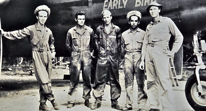Five men in worn, oil-stained coveralls and fatigues stand in a line in front of a bomber's nose lettered "EARLY BIRD", the last letter clipped by the top edge of the print; three wear soft fatigue caps, one a broad-brimmed bush hat and one is bare-headed, and a mainwheel and strut show at the right.