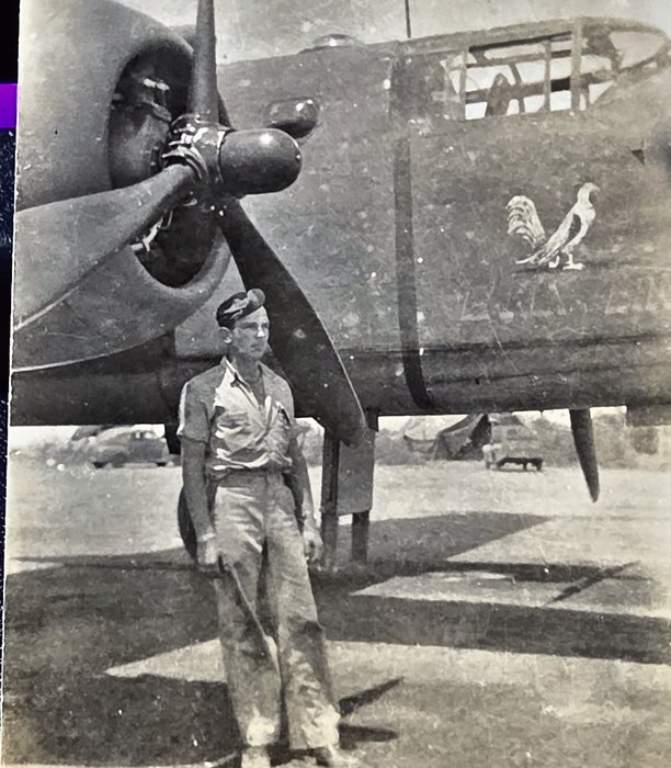 A man in shirtsleeves, trousers and a garrison cap stands beneath the nose of a B-25 Mitchell, a three-bladed propeller above his head and the white rooster of EARLY BIRD on the starboard nose. The name itself is not legible in this frame; the aircraft is identified from the bird. Vehicles, tentage and stacked gear stand on the open field behind him.