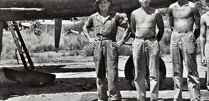 Four men line up beneath an aircraft wing, the man at left in coveralls and a sun helmet with his hands on his hips and the three beside him bare-chested in work trousers and belts. A maintenance ladder is propped against the wing and a main wheel stands behind them; the frame is cropped so that two of the heads are cut off at the top.