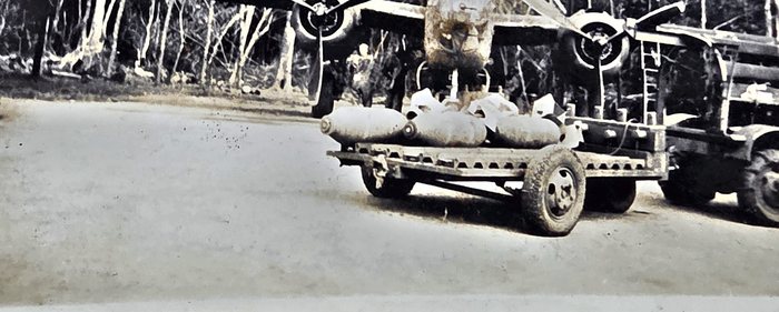 A low four-wheeled bomb trailer, its slotted bed loaded with finned bombs, stands on a bare strip, coupled to a second trailer of the same pattern at right with a vehicle's wheels beyond. Behind it are the mainwheels and undercarriage of a large aircraft, one figure on foot at the treeline.