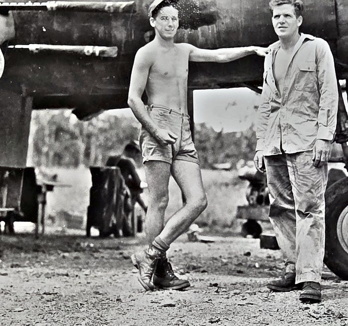 Two men stand beside an aircraft: one bare-chested in cut-down shorts, socks and laced boots with a soft cap, his arm stretched out and his hand resting on the other man's shoulder, the other in a long-sleeved work shirt and trousers. A main wheel is at right and stacked equipment lies in the shade behind them.