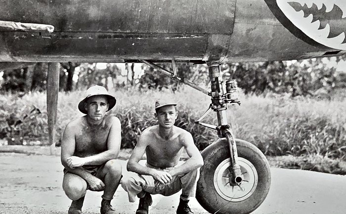 Two bare-chested men crouch on the strip beside a main wheel and its oleo strut under an aircraft wing, one in a pith helmet and one in a fatigue cap. Above and to the right, a row of painted white teeth runs along the fuselage, part of a shark-mouth marking.