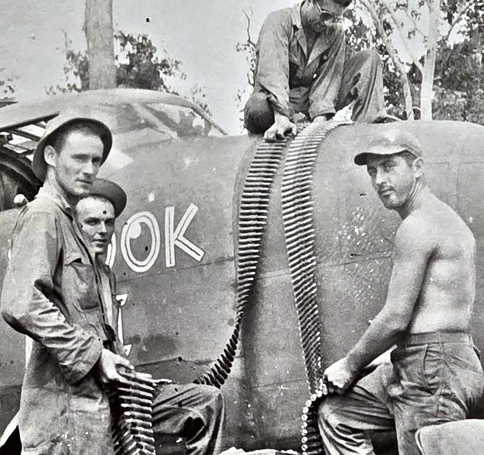 The same photograph as the turret-loading frame in the ground crew section, re-shot from the print. Four men handle long belts of machine-gun ammunition at the fuselage of a B-25, two belts hanging in loops over the top from a man kneeling up there with a steel helmet beside him. The letters <b>OOK</b> stand on the skin in solid white capitals -- the back end of SPOOK, the rest of the name forward of the frame.