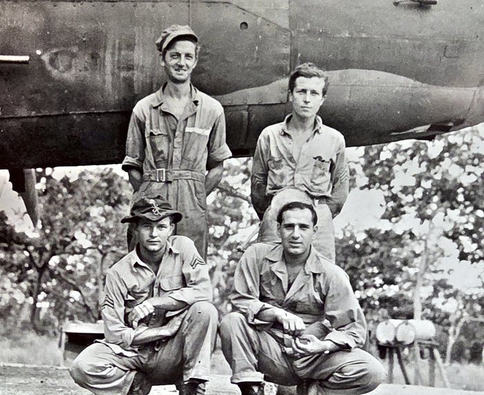 Four men pose in front of an aircraft, two standing -- one in coveralls, one in a work shirt and trousers -- and two crouching in front; the man at lower left wears a broad-brimmed slouch hat with a hat badge and three chevrons on his sleeve. Drums sit on a timber rack at right and a bench stands in the shade beneath the fuselage behind.