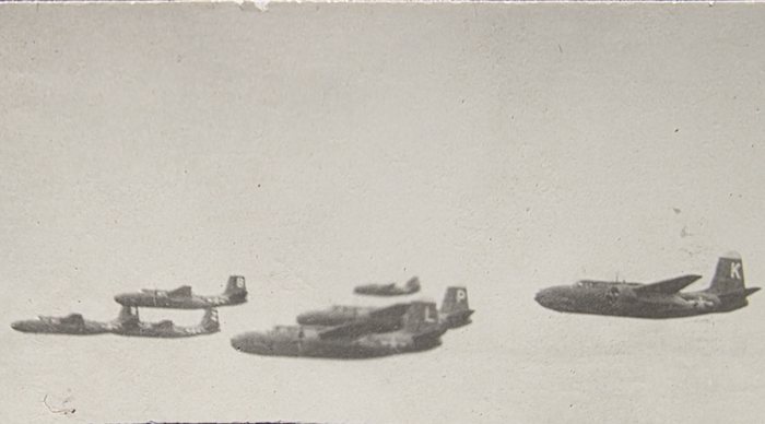 A loose formation of A-20 Havocs, photographed from another aircraft at height against an empty sky. At least five machines are in the frame, strung out in line abreast and stepped down, each carrying a single white letter on the fin -- K on the nearest, P on the one in the centre. The print is a small contact print, faded and torn along the top edge.
