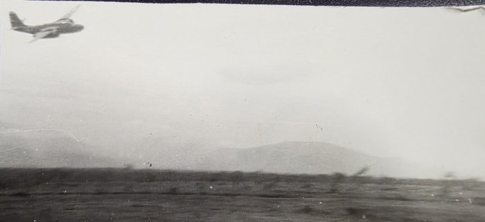 A twin-engine bomber banks away at the top left of the frame while the camera aircraft runs low over a coastline. Dark smoke stands in two or three columns along the shore in the middle distance, with a ridge behind it flattened by haze. The exposure is heavily burnt out in the sky and the print does not say where this is.