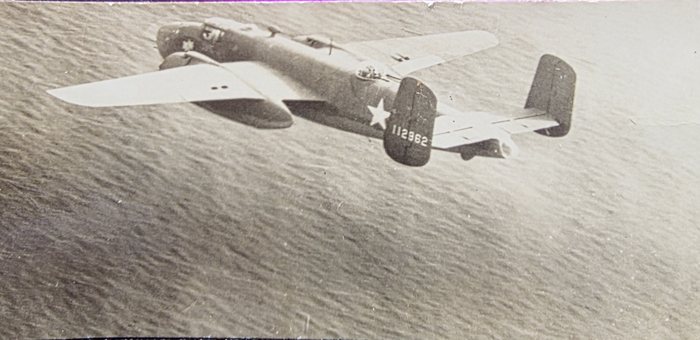 A B-25 Mitchell photographed from above and behind, flying low over open water; the wind ripple on the sea fills the whole background. The twin fins, the gull-wing break outboard of the nacelles and the retracted ventral turret under the rear fuselage are all clear, and the serial 112962 is painted in white on the outer face of the near fin. A white star without bars is carried on the rear fuselage ahead of the tailplane.
