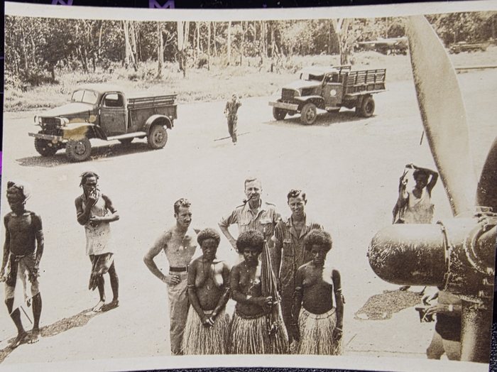 One wide print, not two. A dispersal road runs across the upper half with a Dodge four-wheel-drive pickup at the left, a GMC cargo truck with slatted sides at the right and a man walking between them; in the foreground three American servicemen stand with a group of New Guinean people, four of the women in fibre skirts, while a propeller blade and a large cylindrical store fill the right of the frame and another man reaches up to the aircraft behind them.