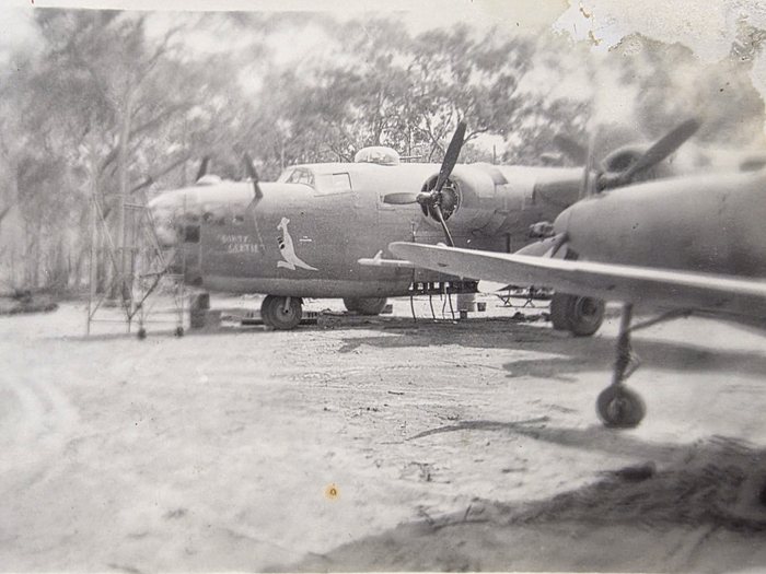 A four-engined bomber standing on a cleared dispersal, photographed from off the port bow. A white kangaroo is painted on the nose with the aircraft's name in two lines of small white capitals beside it; the print is too coarse to read the name with certainty. A maintenance stand stands against the nose, and the wing, engine nacelle and undercarriage leg of a second aircraft fill the right of the frame.
