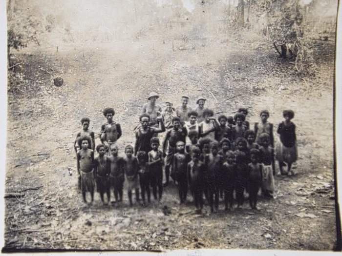 About thirty New Guinean villagers gathered for the camera on cleared, burnt-over ground, with four servicemen standing behind them. The children make up the front two rows; several of the adults behind hold upright staffs. A woman stands a little apart at the right in a dark upper garment and a paler skirt.