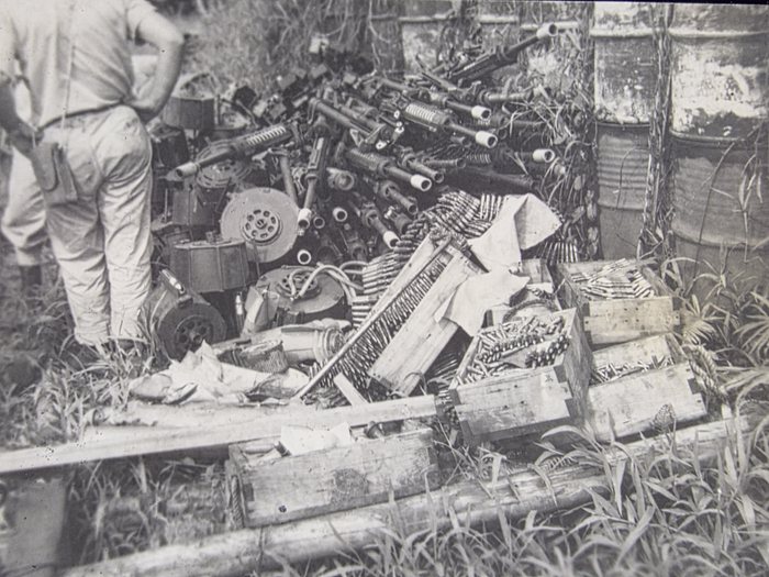 A serviceman in light khaki, his back to the camera and a flap pouch on his belt, stands over a heaped dump of captured Japanese weapons: machine guns with ribbed and perforated barrel jackets, bipods folded beneath them, flared muzzle cones and top-mounted feed housings, piled shoulder-high at the back. Around the foot of the pile lie large flat drum magazines pierced with a ring of holes, faceted metal ammunition canisters, loops of belted ammunition and light wooden crates -- some packed with loose cartridges, one stencilled with a single character that appears to be Japanese. Empty fuel drums stand in the grass behind.