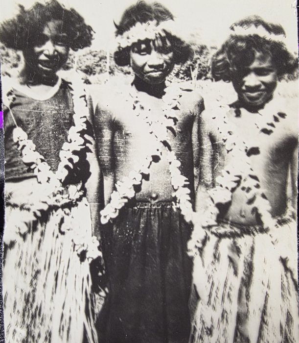 Three young women stand close together facing the camera, low scrub behind them. The two at centre and right wear long garlands of pale flowers over the shoulders and skirts of hanging fibre and are bare above the waist, the centre one with a band of flowers set across her hair; the woman at left wears a dark patterned shirt with her garland over it. All three are smiling. The print is a small one, worn at the edges and scratched across the emulsion.