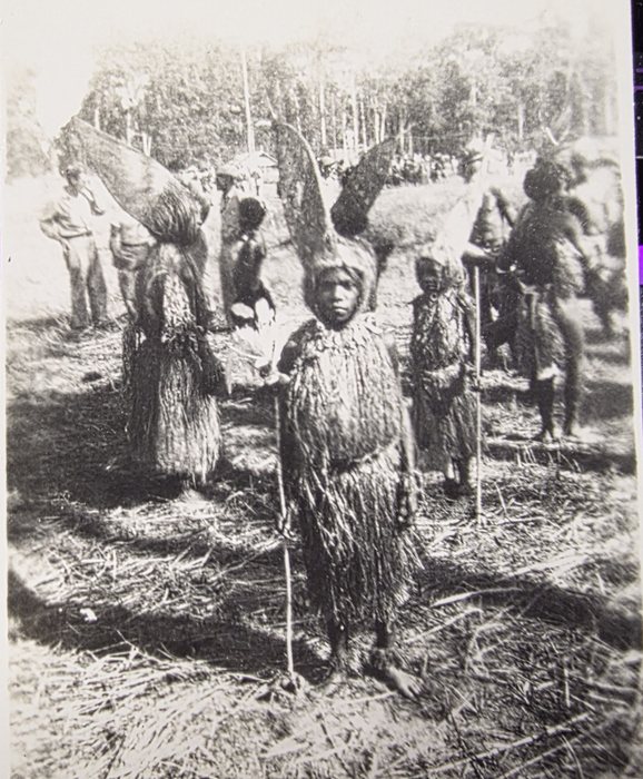 A gathering on cleared, trampled ground with tall thin trees behind: twenty or more people in long cloaks and skirts of hanging fibre, several of them carrying bundles taller than themselves -- broad, curved and tapering to a point, possibly rolled matting or sheets of bark -- up on the shoulder. A child of about ten stands square to the camera in the foreground in a fibre cape and skirt, holding a tall thin staff, with others holding staffs behind. Two men in light shirts and trousers stand watching at the left, and the crowd runs away up the rise to the tree line. The print does not say where this is or what the occasion was.