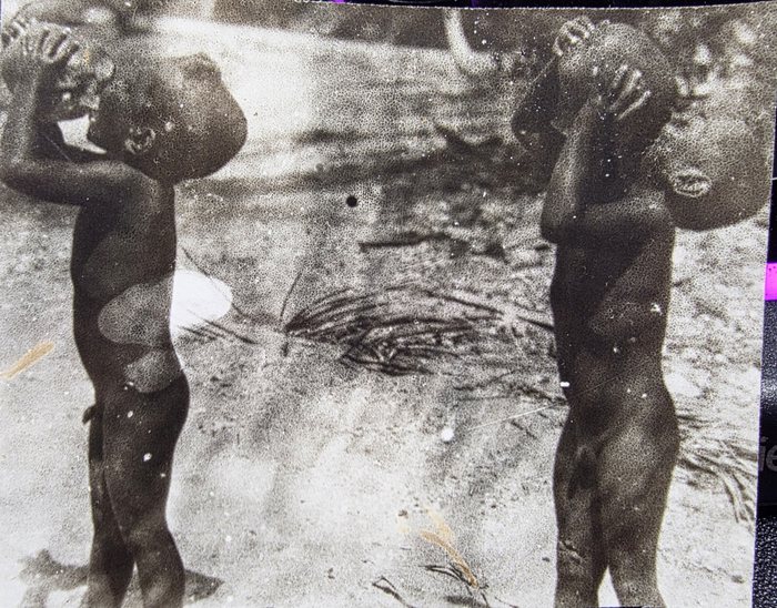 Two children stand on pale sand, each with both arms raised to steady a large rounded load at the head and shoulder; the copy print is too coarse to say what the loads are. A fan of palm frond and a scuffed hollow in the sand lie between them. Neither child is clothed.