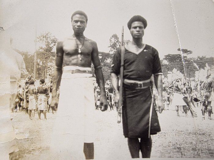 Two men stand facing the camera at the edge of a crowd. The one on the left is bare-chested in a white wrapped cloth and belt, with shell bands on both upper arms and a long pendant on a cord; the one on the right wears a dark long-sleeved jersey over a dark wrap, a broad belt and shell bands at both wrists, and holds an upright wooden shaft in one hand and a thin cane in the other. Behind them stand dozens of people in tall feather headdresses, several carrying hourglass drums.