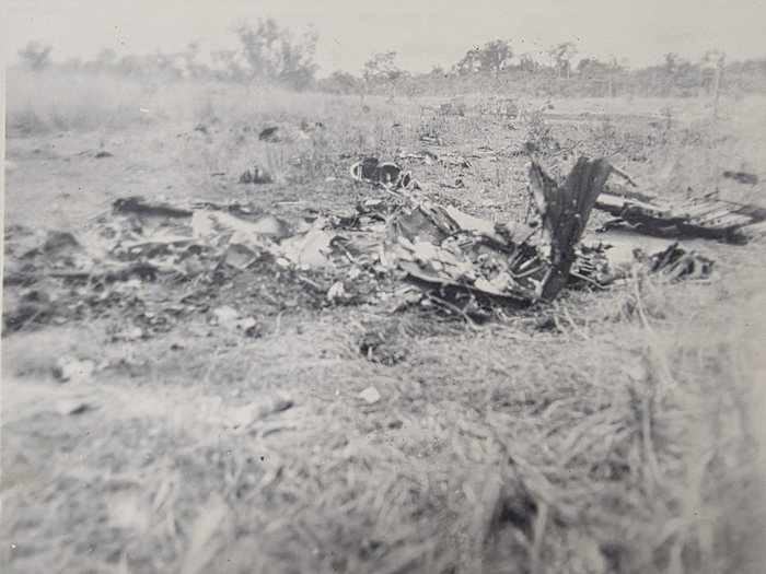 A burnt-out aircraft lying in open, scorched grass. The wreck has broken up and been largely consumed by fire: a dark slab of structure, probably a fin or a section of wing standing on edge, rises at the centre, with a flat ribbed skin panel to the right and what may be an engine mounting ring among the debris at the left. Vehicles or tents are visible on the far edge of the clearing.