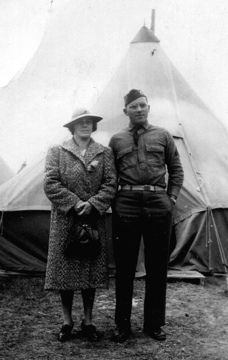 Allen&rsquo;s mother, in a tweed overcoat, light brimmed hat and dark buckled shoes with a handbag held in both hands, standing beside him at the Guard camp &mdash; he is in wool service shirt, tie and garrison cap with a cap insignia, wearing wire-rimmed spectacles. Behind them are pyramidal tents, one with a stovepipe through the roof, and a wooden platform laid at the tent's edge.