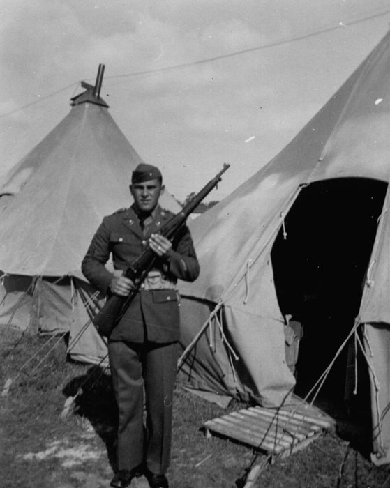 Allen in a wool service coat with collar disks, shirt and tie and a garrison cap, holding a .30-calibre bolt-action rifle across his chest. He stands between pyramidal tents, one with its flap open and a wooden duckboard laid at the entrance.
