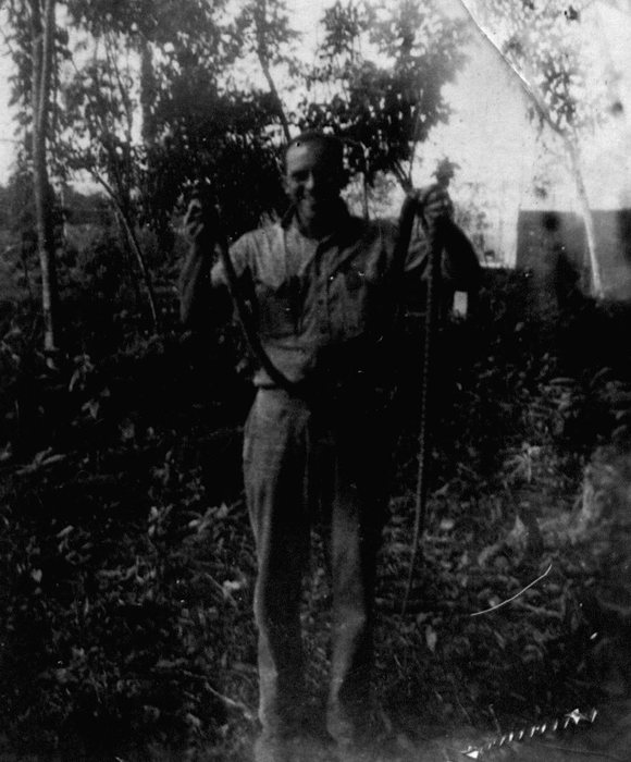 Allen, in shirtsleeves in the low jungle undergrowth, holding up a snake long enough to loop down to the ground beside him &mdash; the pale corner of a tent showing through the trees behind. Identified by the family.