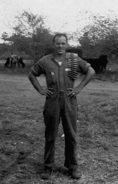 Allen with a belt of machine-gun ammunition draped over his shoulder, cattle grazing behind him.