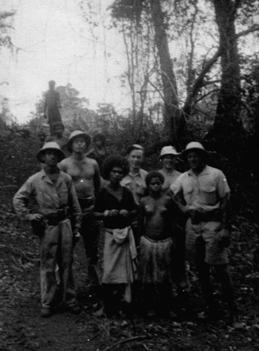 Five servicemen -- four in sun helmets, one stripped to the waist -- stand for a group photograph on a jungle slope with a New Guinean woman and a girl, who are in the front rank facing the camera: the woman in a dark wrap with a pale cloth apron and her hands held together in front of her, the girl beside her bare above the waist in a skirt of hanging fibre. A further head shows at the back of the group, too indistinct to count either way, and a lone figure is silhouetted on the rise behind.