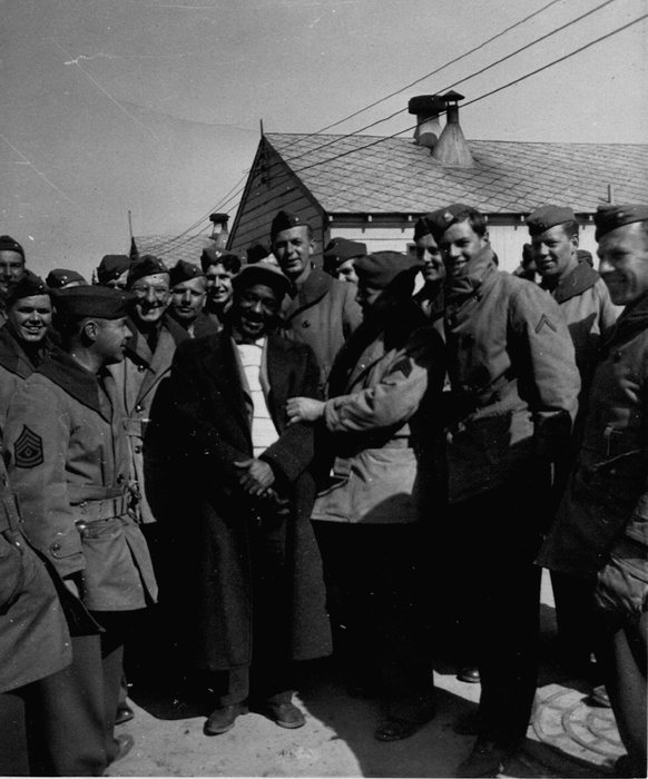 A crowd of soldiers in wool service coats and garrison caps presses in around a Black civilian in a long overcoat, striped sweater and soft cap, who is laughing as he shakes hands with one of them. Wooden barracks with shingled roofs and metal roof ventilators stand behind; a first sergeant's chevrons show on the sleeve at the left of the frame.